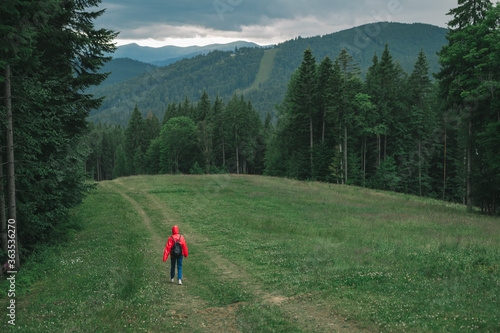 Minimalistic photo, back view on tourist woman in red raincoat walking on path, mountain chain on background. Greenery landscape, full-length picture of traveler girl, strolling among the mountains.