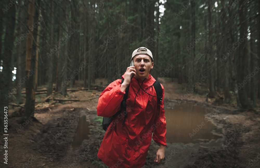 © bodnarphoto - The terrified male traveler is talking on the phone, shouting in the mountain fir forest. A young tourist man in a red jacket with a backpack is feeling scared and lost in the woods.