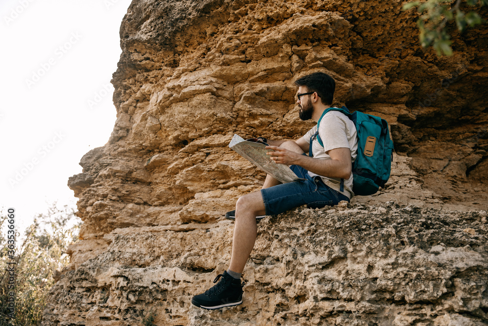 Young man sitting on a rock in mountains, holding a map, planning routes.