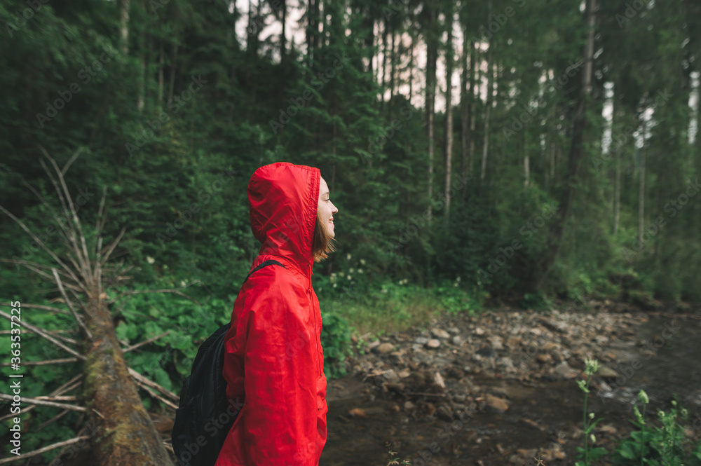 Side view on a cheerful girl-traveler looking at the mountain river and smiling. Joyful young female wearing a red hood and raincoat, feeling happy in the mountain forest, looking far ahead.