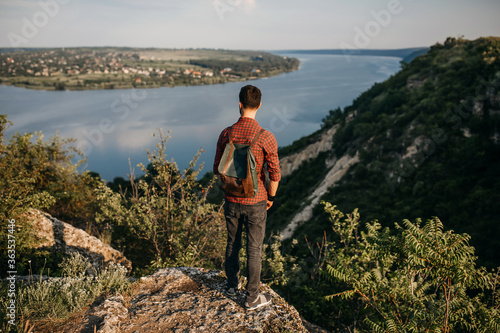 Young bearded man with a leather backpack standing on a cliff in mountains, looking towards a river. Concept of travel. © Bostan Natalia
