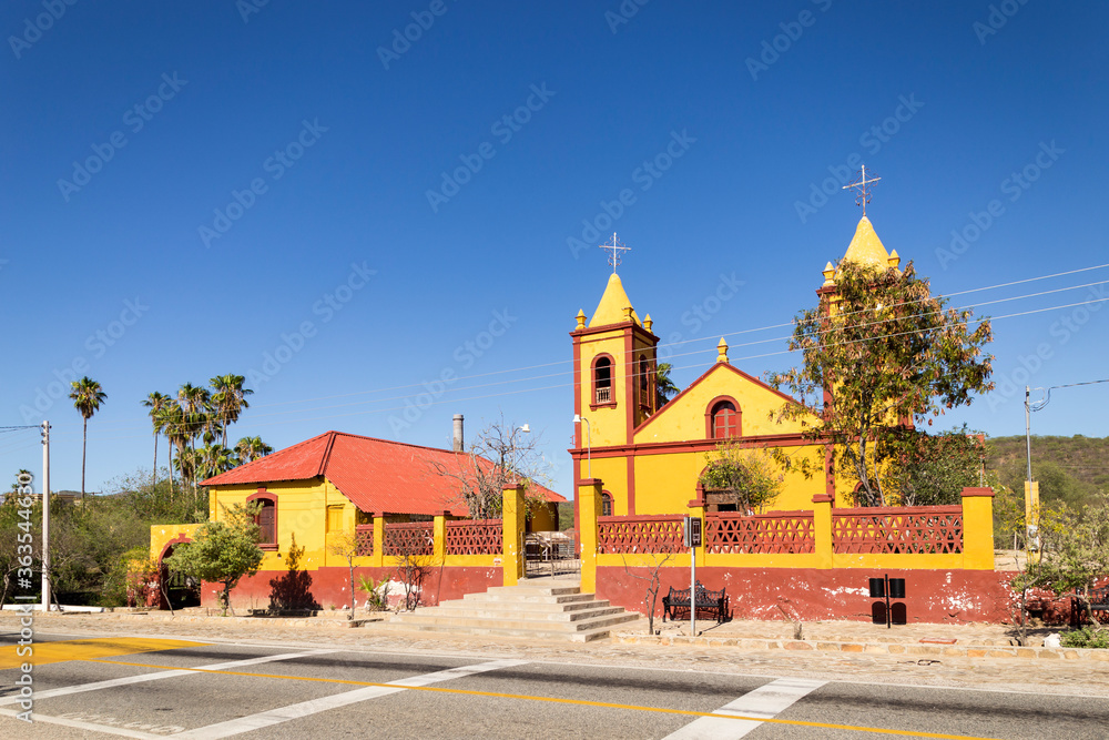 Horizontal wide view of empty road and small yellow and red colored Mexican Our Lady of Guadalupe parish under clear blue sky, in former mining town of El Triunfo, Baja California Sur, Mexico