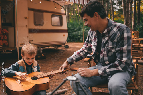 Father teaches son play guitar on camping trip relaxing in the autumn forest. Camper trailer. Fall season outdoors trip