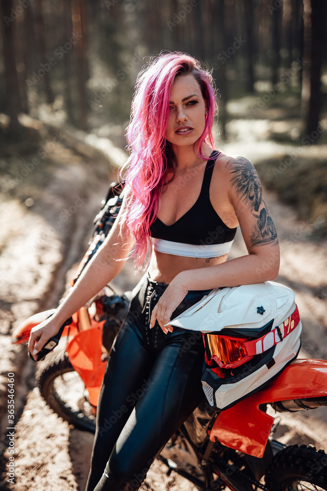 Beautiful tattooed hipster girl with bright pink hair sitting on her motocross bike in woods