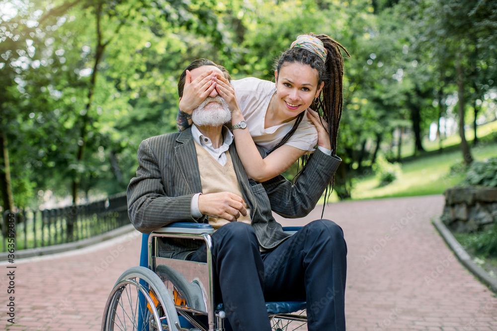 Young smiling Caucasian woman with dreadlock hair covering the eyes of her senior disabled grandfather in wheelchair, making surprise for him on a walk in the park