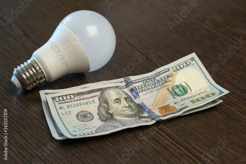 A light bulb and hundred-dollar bills lies on a brown wooden table. Electricity payment concept. Close up.