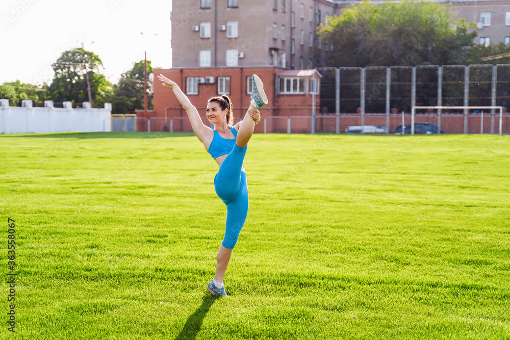 Overall plan. Beautiful brunette girl athlete, dressed in a top and tight leggings, doing sports exercises on a city field. Raising leg up, casting a shadow on the grass. Stretching, training her body