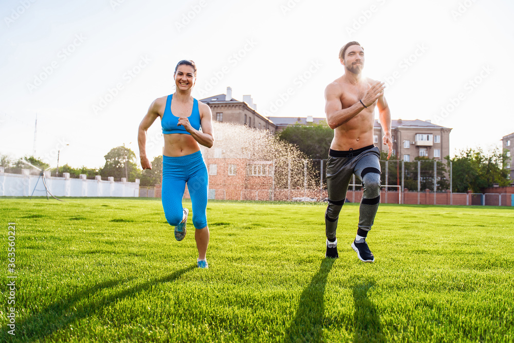 Sportsmen man and woman running in stadium. Healthy sport activity for adults. Young athletes in training, runner exercising. Dynamic run of sprinters in a city stadium on the grass Sportsmen man and woman running in stadium. Healthy sport activity for adults. Young athletes in training, runner exercising. Dynamic run of sprinters in a city stadium on the grass