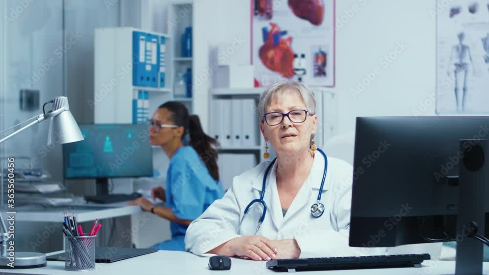 Authentic portrait of an experienced senior doctor smiling at the camera with the nurse in background working. Modern private hospital consultation room doctor cabinet Family therapist Authentic portrait of an experienced senior doctor smiling at the camera with the nurse in background working. Modern private hospital consultation room doctor cabinet Family therapist