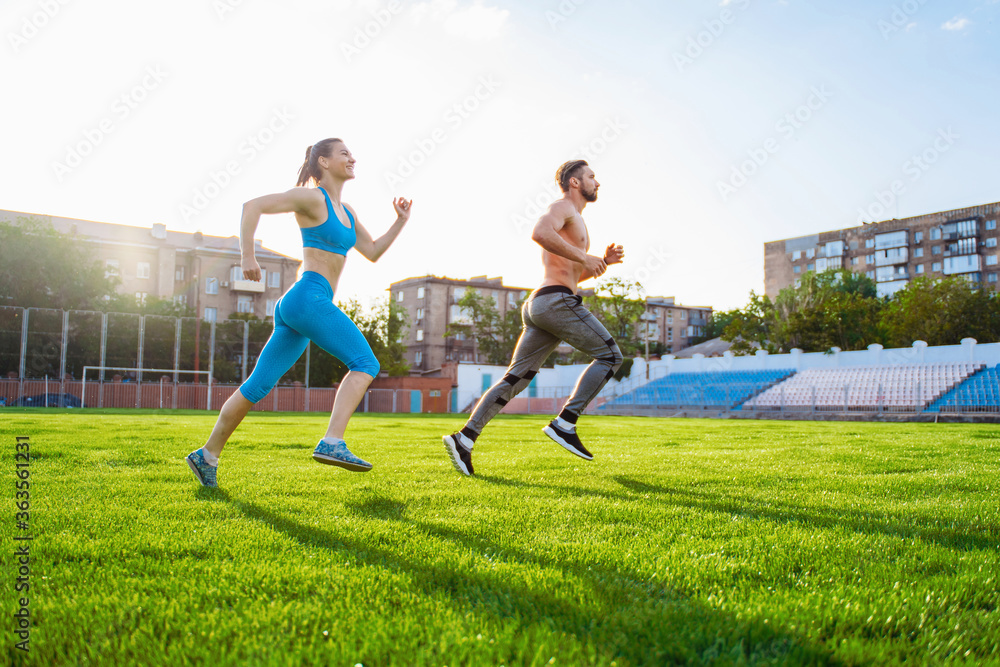 Sportsmen man and woman running in stadium. Healthy sport activity for adults. Young athletes in training, runner exercising. Dynamic run of sprinters in a city stadium on the grass