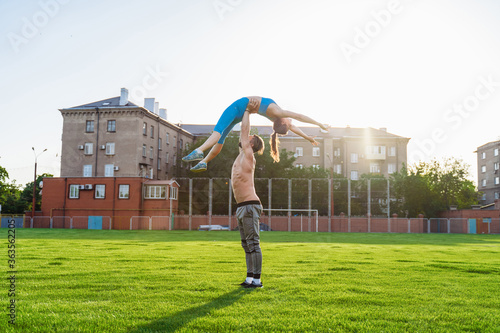 Fit young couple doing acro yoga in the park or on the field stadium. Man standing on the grass and balancing his woman on hands. Happy, sportive young athletes
