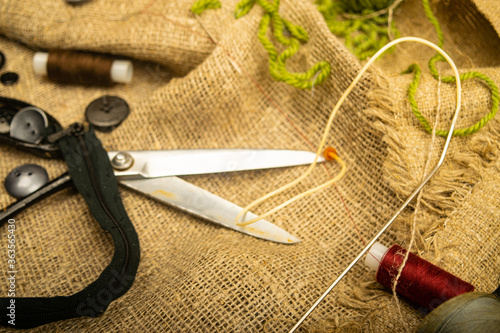 Buttons of various sizes, a spool of thread, tailor's scissors, and a zipper on rough-textured burlap. Close up.