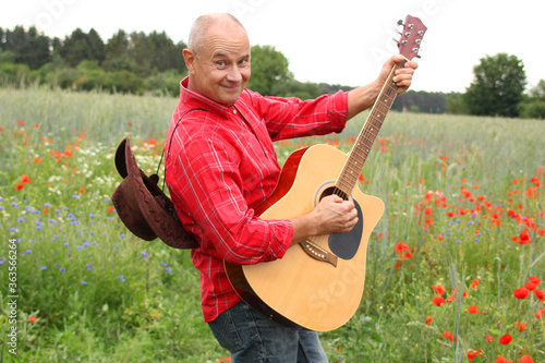 cheerful adult man in a red shirt and a coboy hat on a field of green poppies plays guitar, posing and having fun, concept musical, inspiration by nature, beauty of the native land photo