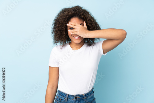 Young African American woman isolated on blue background covering eyes by hands and smiling