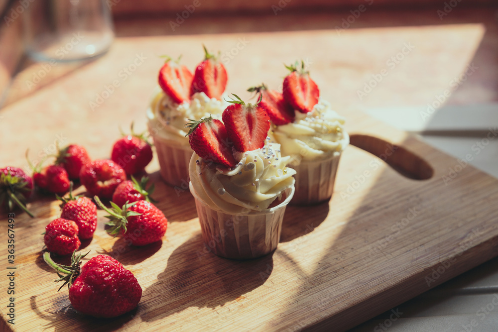 Appetizing cupcakes with white cream decorated with fresh strawberries lit by the rays of the sun. Pleasant drinking. Appetizing cupcakes with white cream decorated with fresh strawberries lit by the rays of the sun. Pleasant drinking.