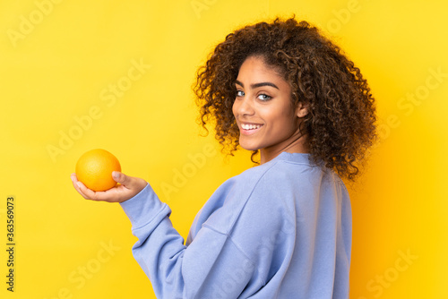 Young African American woman holding an orange over yellow background