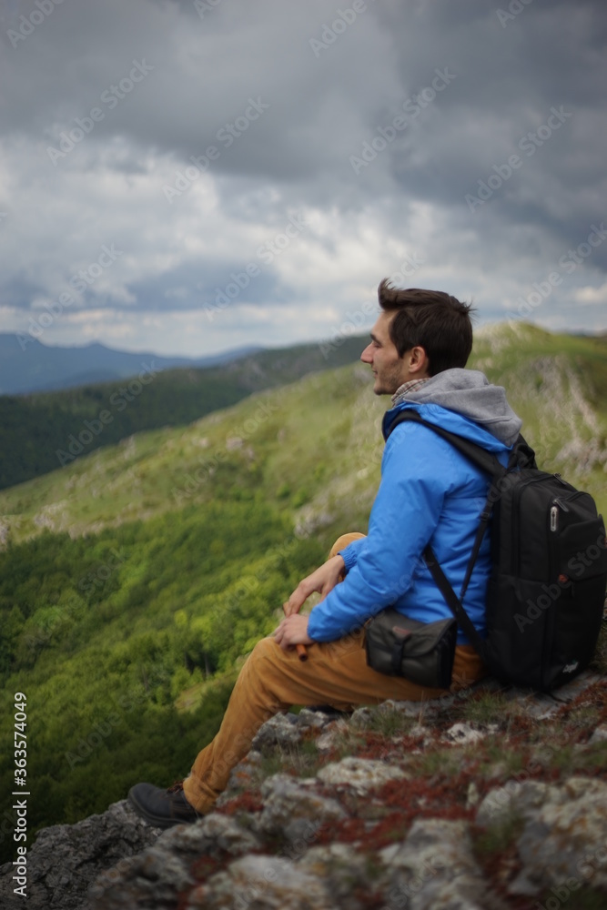 A young male tourist sitting on a rock. He is on the summit of Stara Planina (Balkan Mountains) and enjoying the view. In one of his hands, he's holding a wooden flute/ pipe.