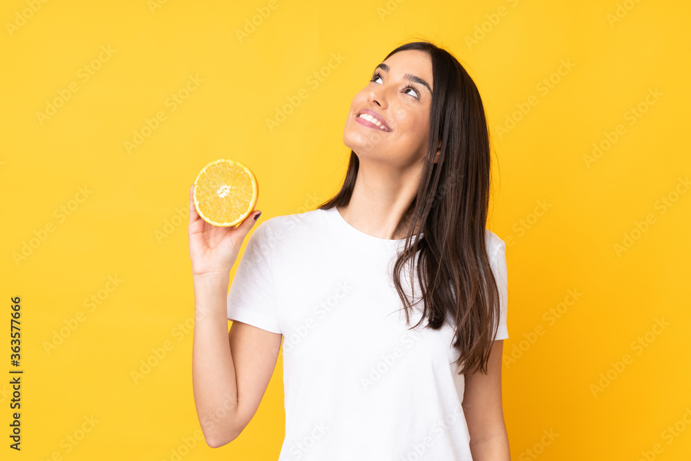 Young caucasian woman holding an orange isolated on yellow background looking up while smiling Young caucasian woman holding an orange isolated on yellow background looking up while smiling