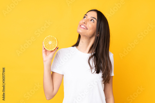 Young caucasian woman holding an orange isolated on yellow background looking up while smiling