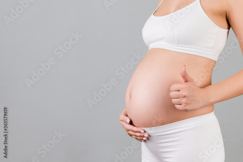 Cropped shot of positive pregnant woman in white underwear showing thumb up cool gesture over her baby bump at gray background. Happy maternity. Copy space