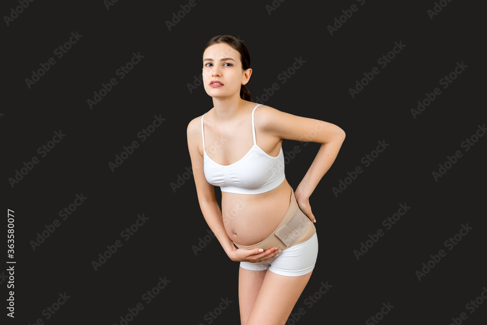 Portrait of supporting bandage against backache on pregnant woman in underwear at black background with copy space. Mother is suffering from pain in the back. Orthopedic abdominal support belt concept Portrait of supporting bandage against backache on pregnant woman in underwear at black background with copy space. Mother is suffering from pain in the back. Orthopedic abdominal support belt concept