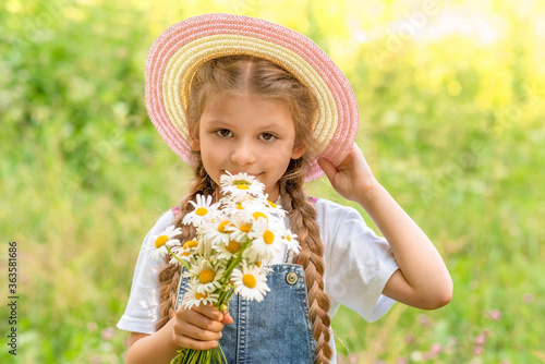 A little girl in a straw hat sniffs a bunch of wild flowers in a green clearing.