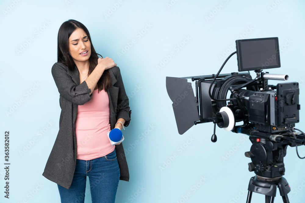Reporter woman holding a microphone and reporting news isolated on blue background suffering from pain in shoulder for having made an effort Reporter woman holding a microphone and reporting news isolated on blue background suffering from pain in shoulder for having made an effort