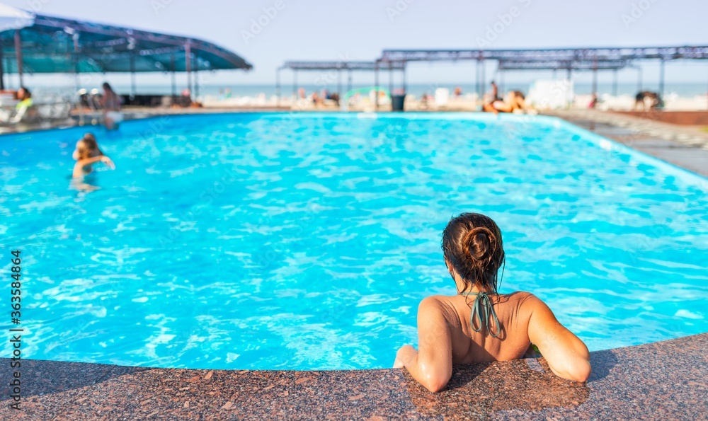 Rear view happy little teenage girl leaned on edge of the pool and enjoys clean warm water under the rays of the hot summer sun while relaxing at the hotel. Sea vacation concept. Place for advertising