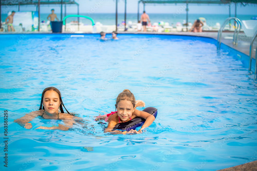 Two little sisters girls are swimming in a large pool with clear blue water of mine near the hotel on the background of the sea and the beach. Concept vacation tropical hot country with children