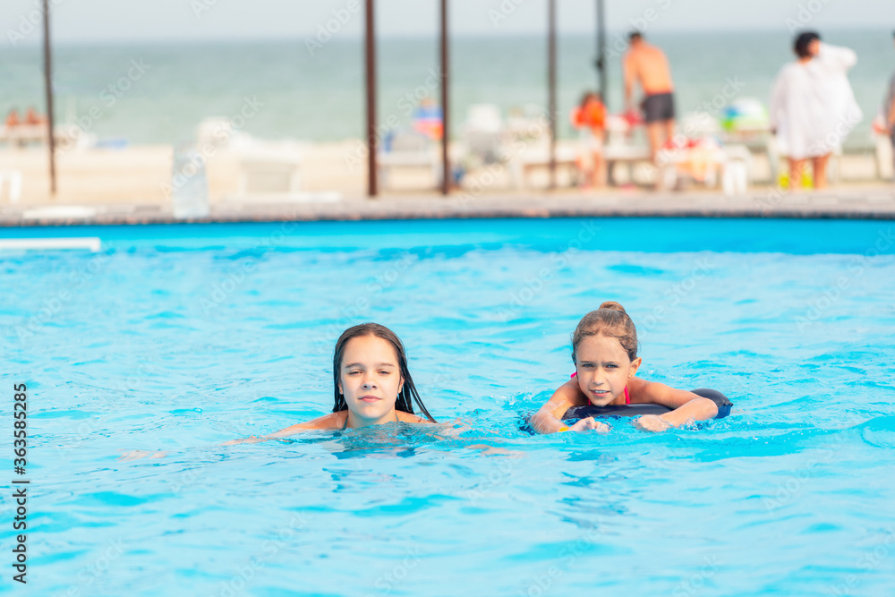 Fototapeta premium Two little sisters girls are swimming in a large pool with clear blue water of mine near the hotel on the background of the sea and the beach. Concept vacation tropical hot country with children