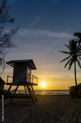 Wooden lifeguard rescue tower on the empty beach with sunset cloudy sky, sea background and a palm tree © OlgaPS