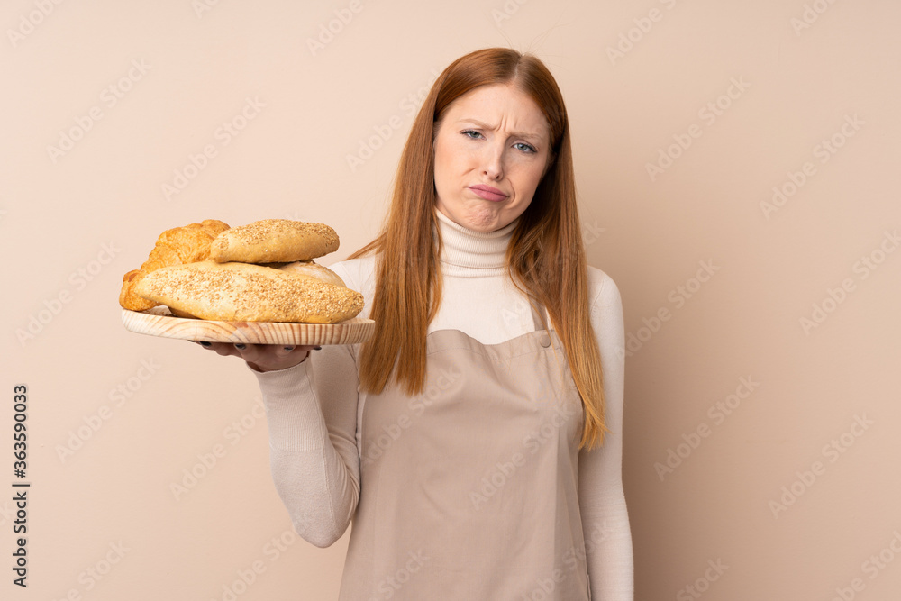 Young redhead woman in chef uniform. Female baker holding a table with several breads with sad expression Young redhead woman in chef uniform. Female baker holding a table with several breads with sad expression