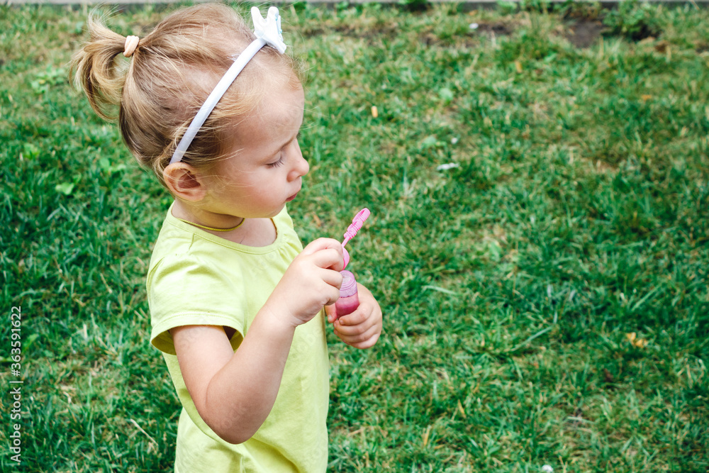 Summer outdoor activities for children. A little blonde girl in a yellow t-shirt blows soap bubbles against the background of green bright grass in the courtyard with a blur of focus. Summer outdoor activities for children. A little blonde girl in a yellow t-shirt blows soap bubbles against the background of green bright grass in the courtyard with a blur of focus.