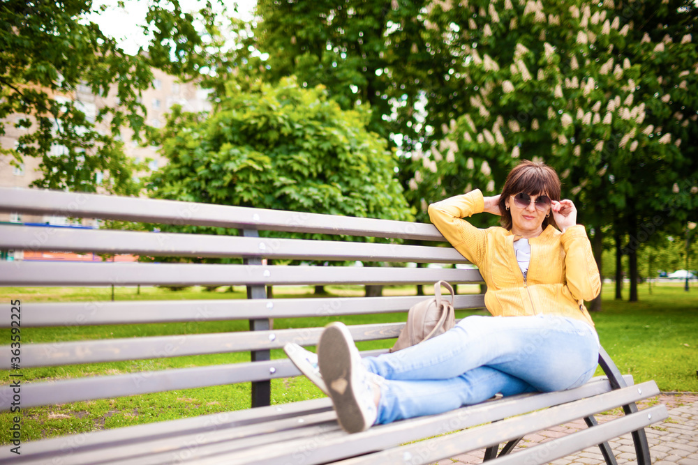 Pretty relaxed well-groomed middle-aged caucasian woman in casual clothes resting sitting on bench in park on sunny warm summer day. Concept of the long-awaited rest after coronovirus Pretty relaxed well-groomed middle-aged caucasian woman in casual clothes resting sitting on bench in park on sunny warm summer day. Concept of the long-awaited rest after coronovirus