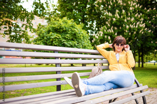 Pretty relaxed well-groomed middle-aged caucasian woman in casual clothes resting sitting on bench in park on sunny warm summer day. Concept of the long-awaited rest after coronovirus