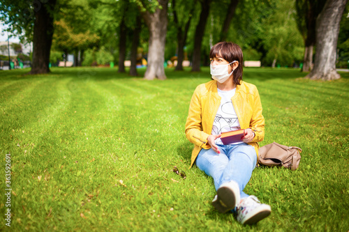 Middle-aged young woman in casual wear puts on a white medical mask sitting on the floor in a summer sunny park. Coronovirus pandemic concept