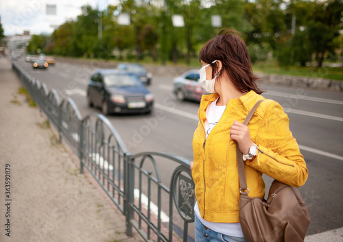 Adult pretty woman in casual clothes and bag with a protective mask on her face is standing by the road with passing cars. Coronovirus protection concept in a megalopolis