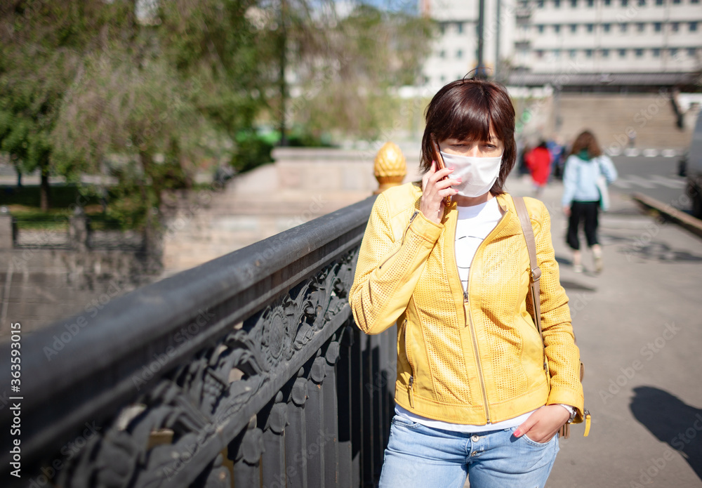 Funny middle-aged woman in a white protective mask talking on a smartphone while walking around the city on a warm spring day. Coronovirus pandemic and remote communication concept
