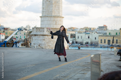 A beautiful girl with long black hair and black coat with red and black striped dress uses your phone while waiting near the pier of the port of Molfetta photo