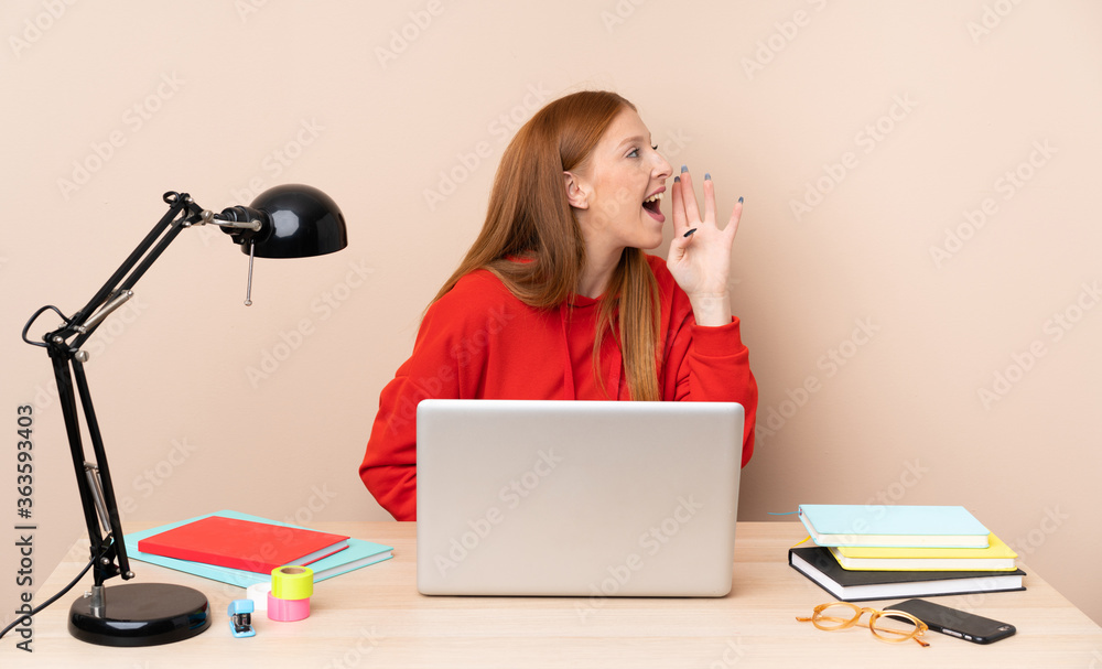 Young student woman in a workplace with a laptop shouting with mouth wide open to the lateral Young student woman in a workplace with a laptop shouting with mouth wide open to the lateral