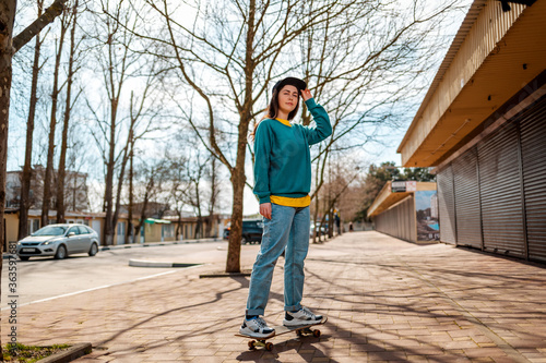 A young Caucasian hipster woman poses standing on a skateboard. In the background, an alley. Concept of sports lifestyle and street culture © _KUBE_