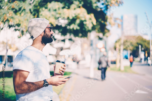 Young good looking bearded blogger in stylish sunglasses enjoying recreation with delicious take away coffee at urban setting.Bearded male person in white hat spending time outside with modern device