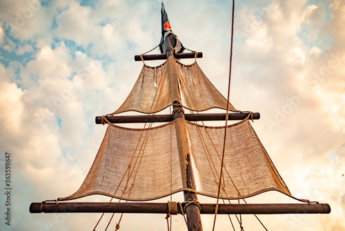 Fényképezés Bottom view of a ship mast with beige sails swings against a blue sky with sunny sunny summer warm day clouds