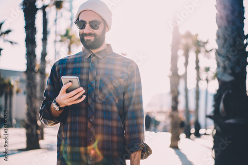 Positive hipster guy in sunglasses and stylish hat installing application on smartphone device walking in urvan setting.Bearded student dressed in casual clothes reading sms message on cellular photo