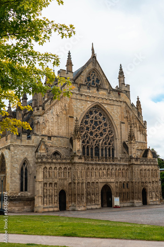 Exeter Cathedral side on image with cloudy sky in the background and framed by green trees and grass in the foreground