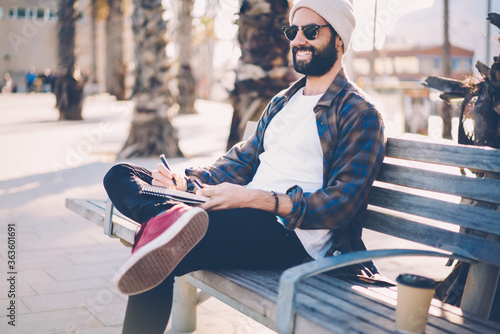 Cheerful bearded guy in sunglasses sitting on bench spending free time on hobby making sketches in notepad.Smiling talanted male painter in hat drawing in notebook enjoying leisure time outdoors photo