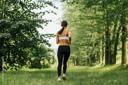 Back view of woman in sportswear jogging on green grass at park. Healthy sport lifestyle. Concept sportive woman. Active female running outdoors.