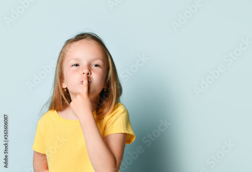 Blonde child in yellow t-shirt. She is showing be quiet sign by her forefinger while posing against turquoise studio background. Close up