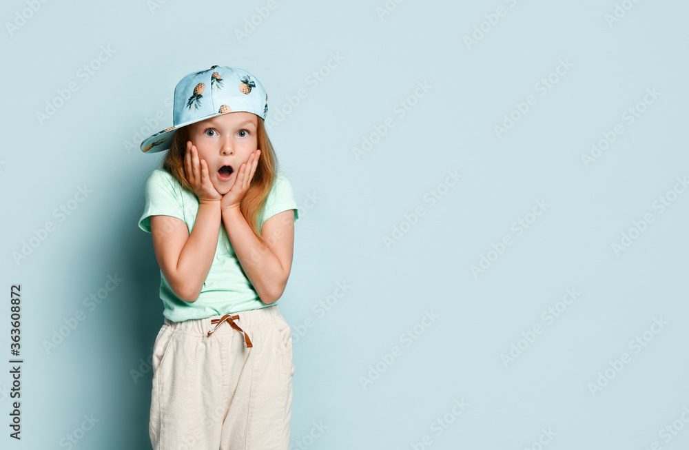 Child in t-shirt, pants and cap printed by pineapples. She raised hands and looking shocked, posing against turquoise background. Close up