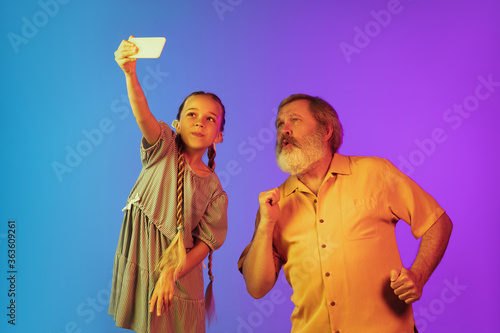 Taking selfie, vlog together. Senior man spending happy time with granddaughter in neon. Joyful elderly lifestyle, family, childhood, tech concept. Enjoying blogging, taking shoots. Copyspace.